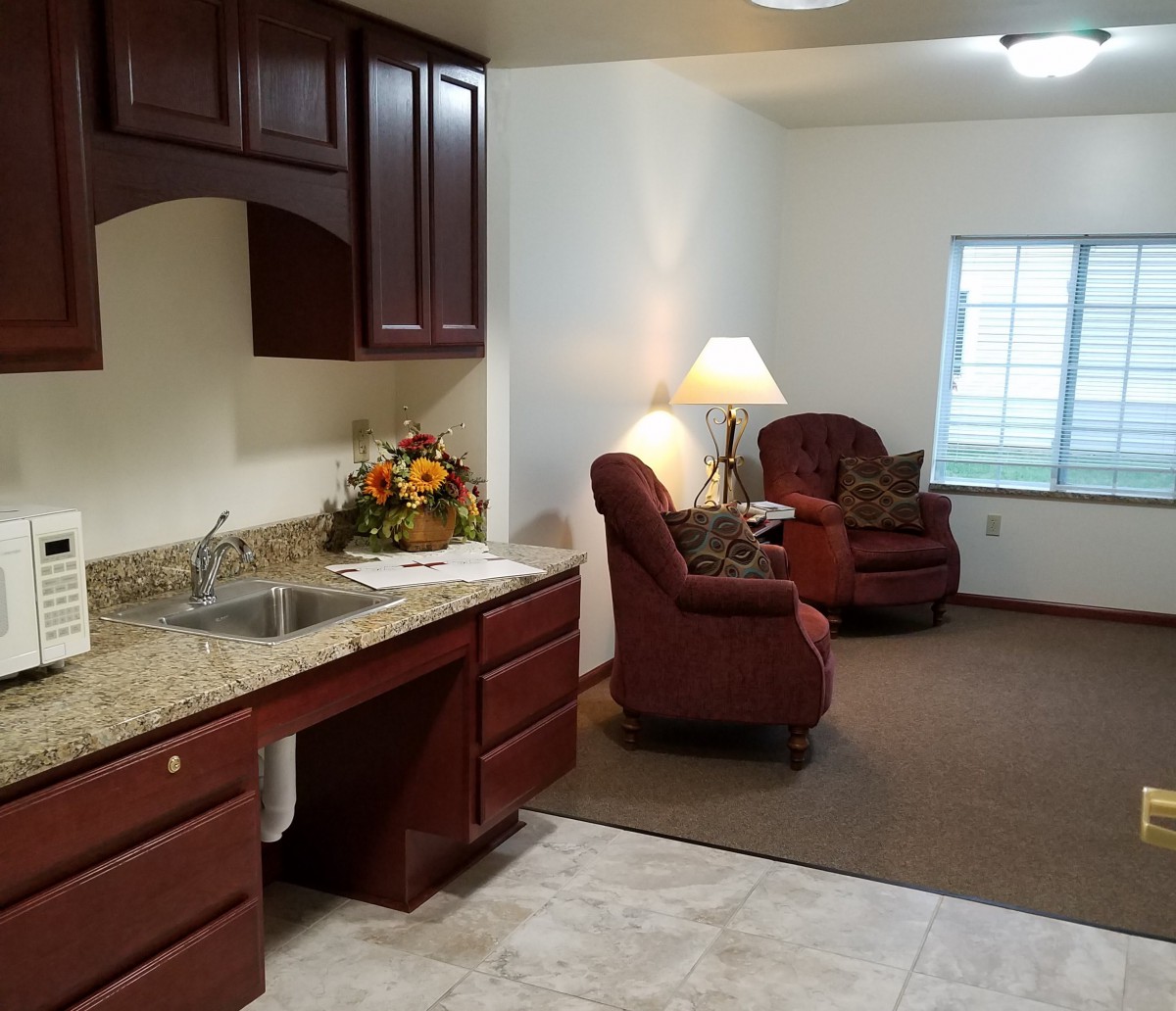 A cozy living area with a small kitchen space, featuring armchairs, a lamp, and cabinets.