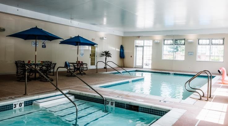 An inviting indoor swimming pool area with a hot tub, surrounded by umbrella-shaded tables and chairs for relaxation.