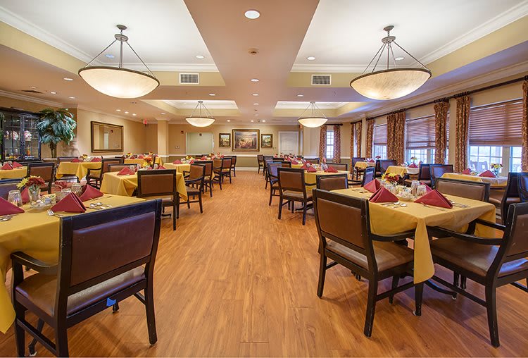 A spacious and elegantly designed dining area with neatly arranged tables and chairs, featuring warm lighting from chandeliers and a wooden floor.
