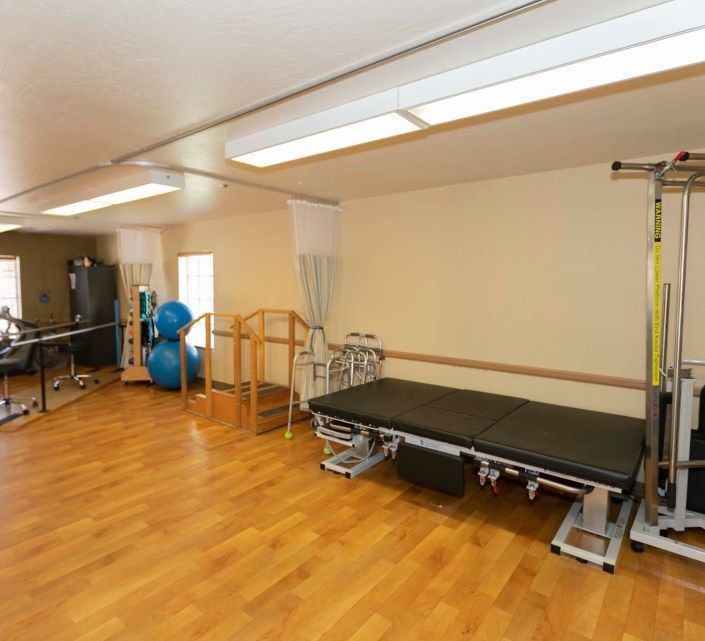 A spacious rehabilitation therapy room equipped with exercise balls, a treatment table, and parallel bars on a wooden floor.