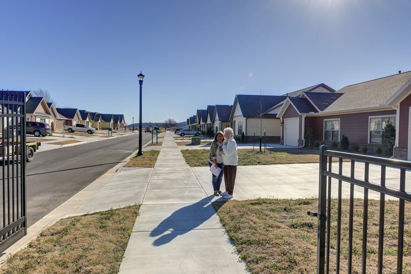 Two people walking on a sidewalk in a suburban neighborhood under a clear blue sky.