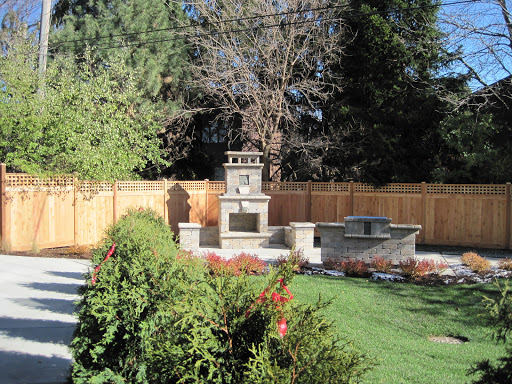 A serene garden featuring a stone fountain, patio area, and lush greenery surrounded by a wooden fence.