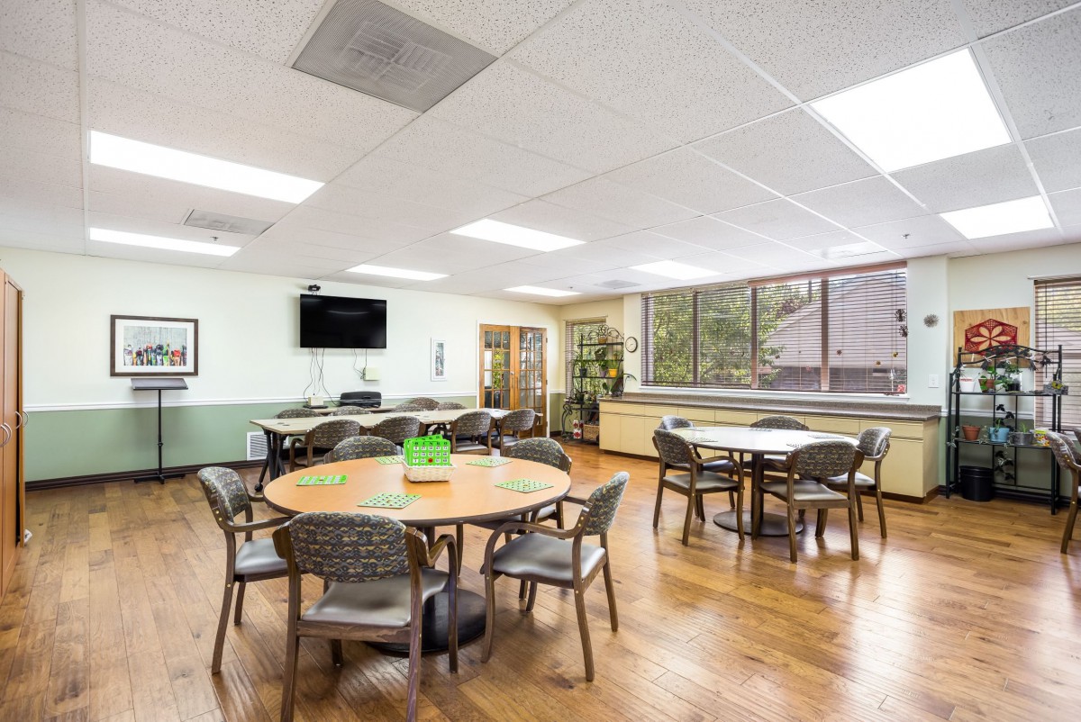 A well-lit room with several tables and chairs arranged, featuring a television on the wall and large windows.