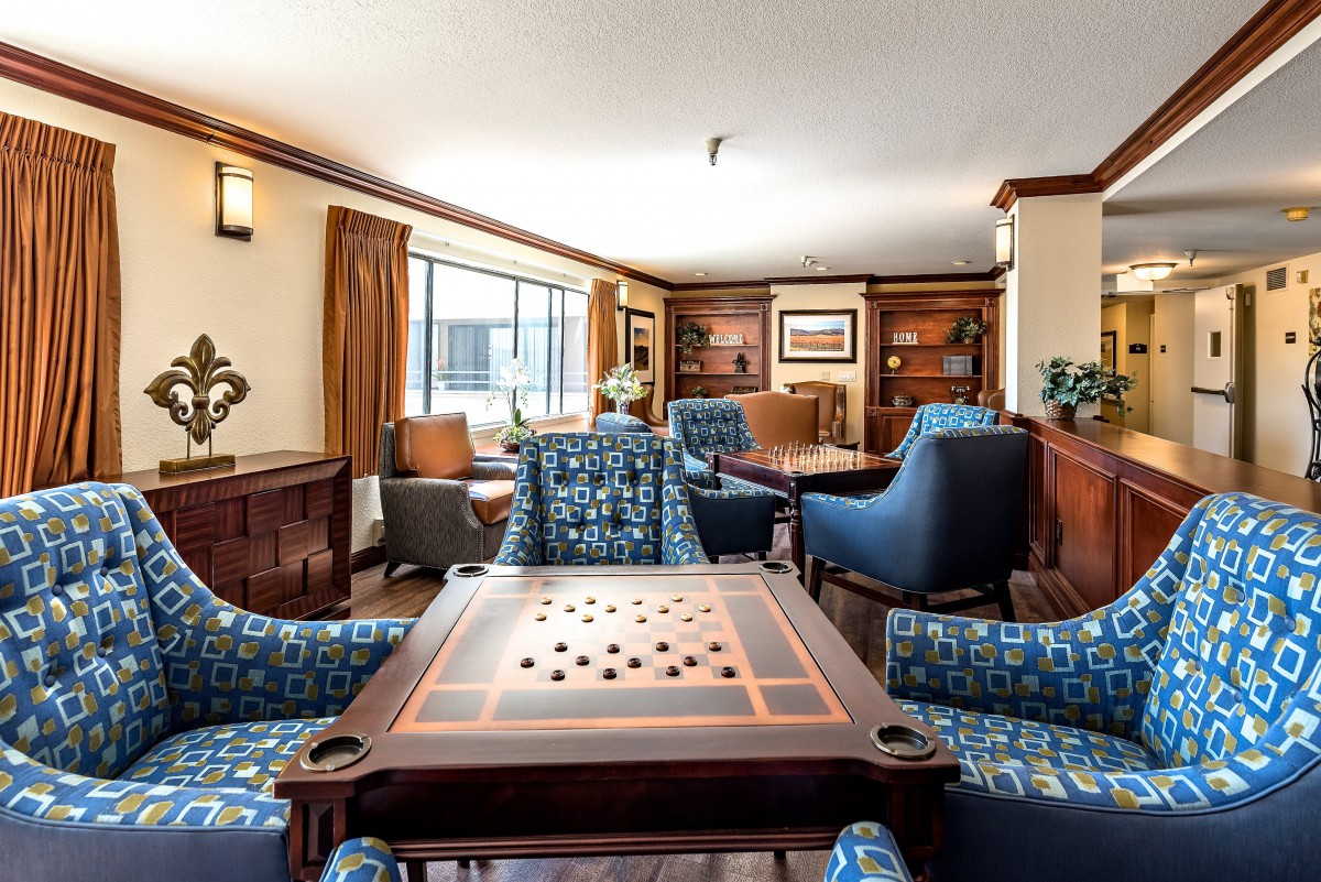 A cozy living room with a game table, surrounded by comfortable chairs and bookshelves, bathed in natural light.