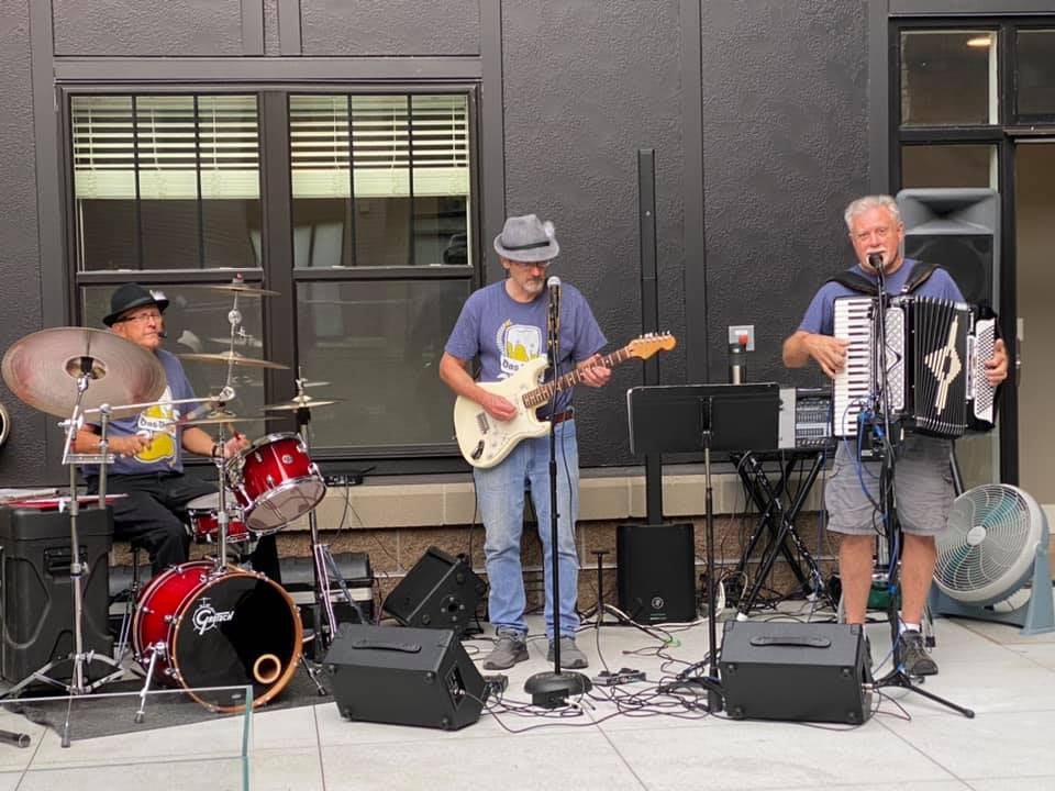 A band performs live outdoors, featuring musicians playing drums, guitar, and accordion.