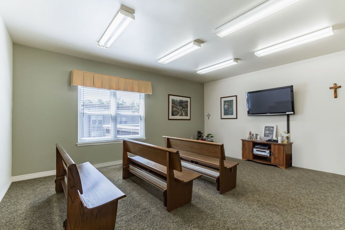 A small room with wooden benches facing a television, featuring windows and ceiling lights.