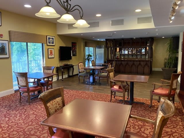A cozy lounge and dining area with several tables and chairs, featuring a carpeted floor, a chandelier, and a television mounted on the wall.