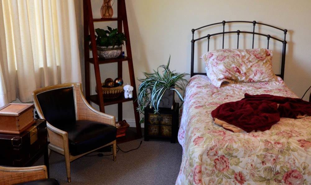 A cozy bedroom with a floral bedspread, an elegant chair, and decorative plants on shelves.