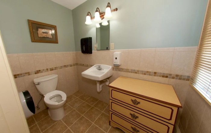 A clean and neatly organized bathroom with a sink, toilet, mirror, and a piece of furniture.