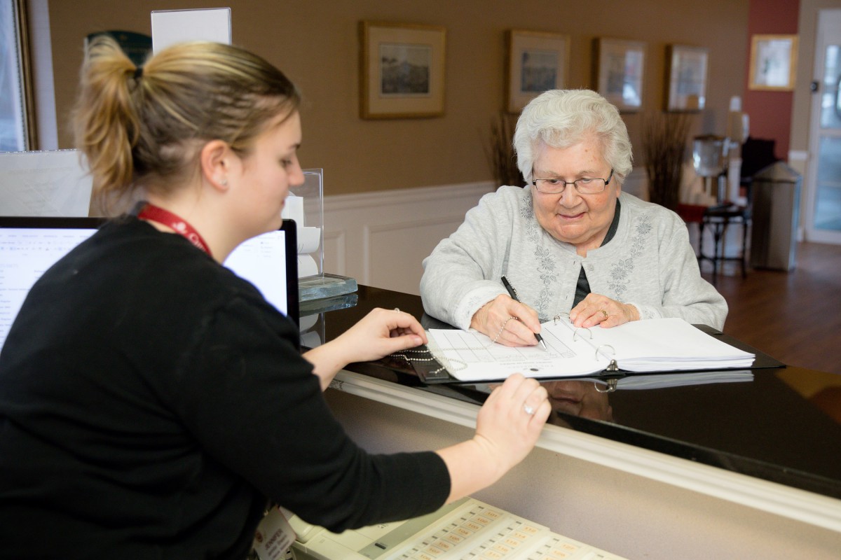 An elderly woman signing documents at a reception desk.