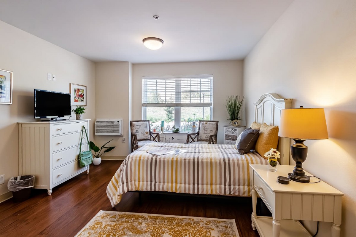 A cozy and well-decorated bedroom featuring a bed with striped bedding, a large window, and various furnishings including a dresser and potted plants.