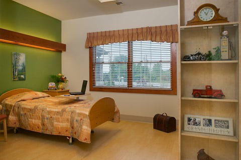 A cozy hospital room featuring a single bed, large window with blinds, and shelves for personal items.
