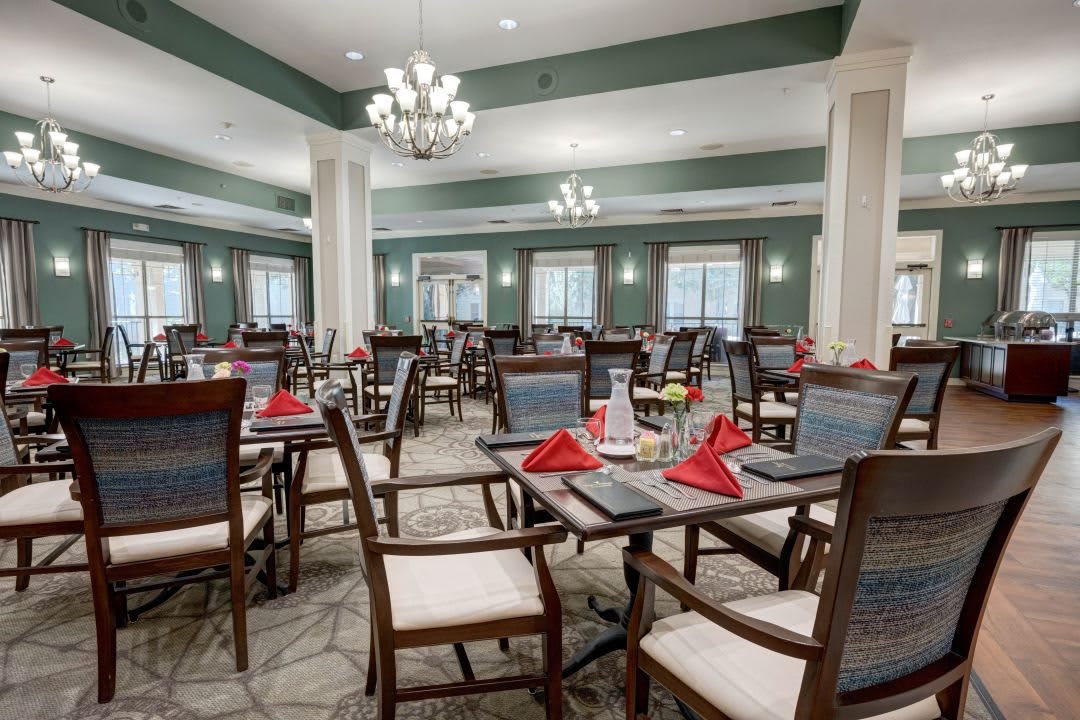 A well-decorated dining room with neatly arranged tables and chairs, featuring red napkins and chandeliers.