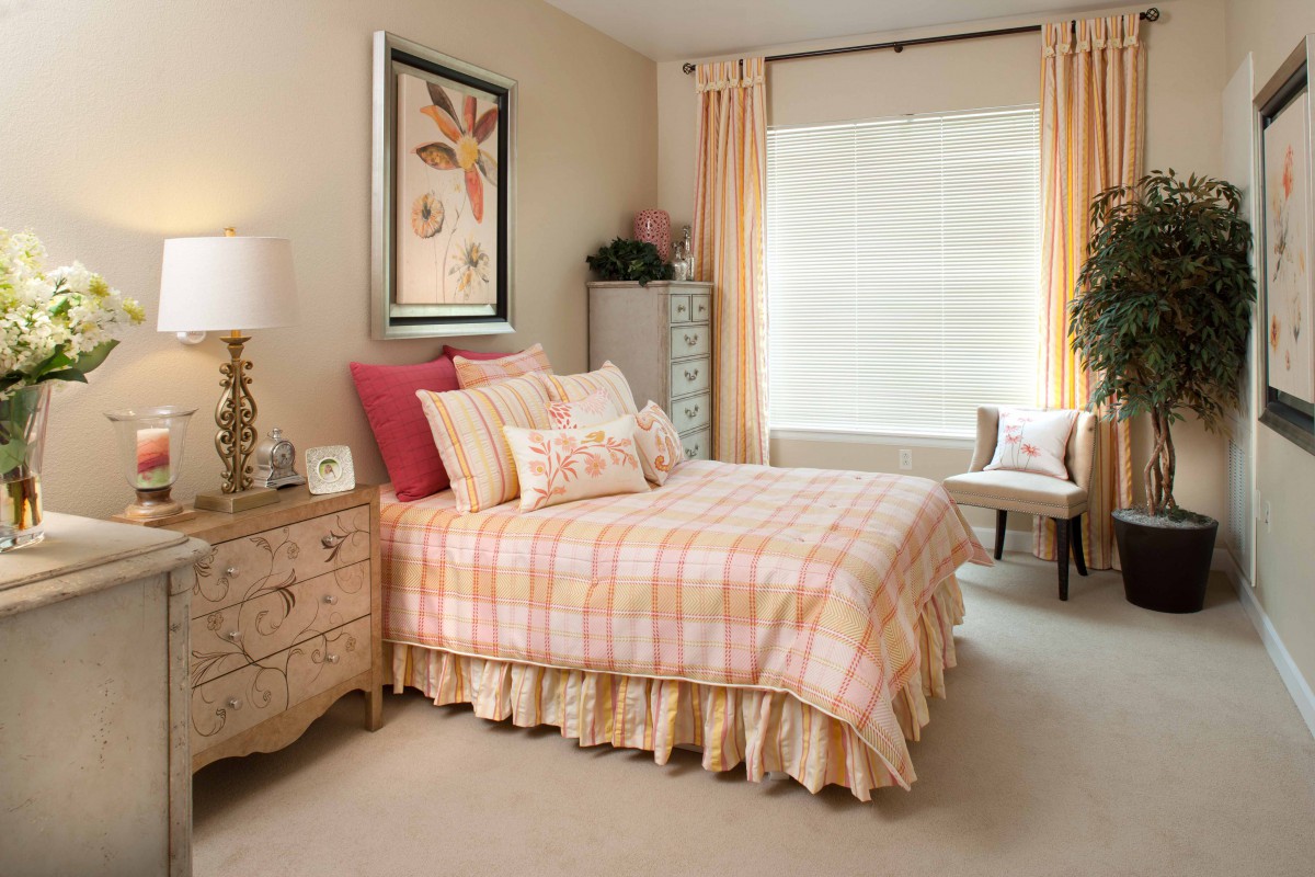 A cozy bedroom with a pink and beige color scheme featuring a neatly made bed, decorative pillows, a nightstand with a lamp, and a window dressed with light-colored curtains.