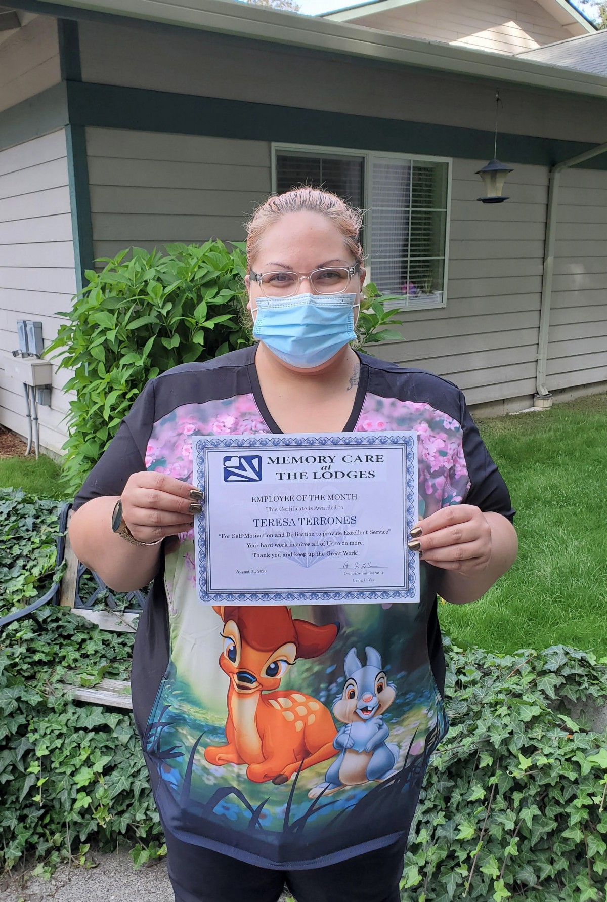 A person wearing a mask and holding a certificate stands outside near greenery.