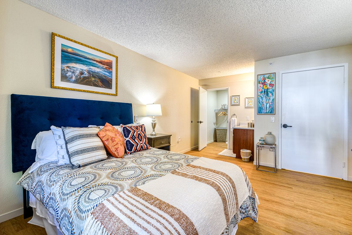 A cozy bedroom with a patterned bedspread, artwork on the wall, and a wooden floor.