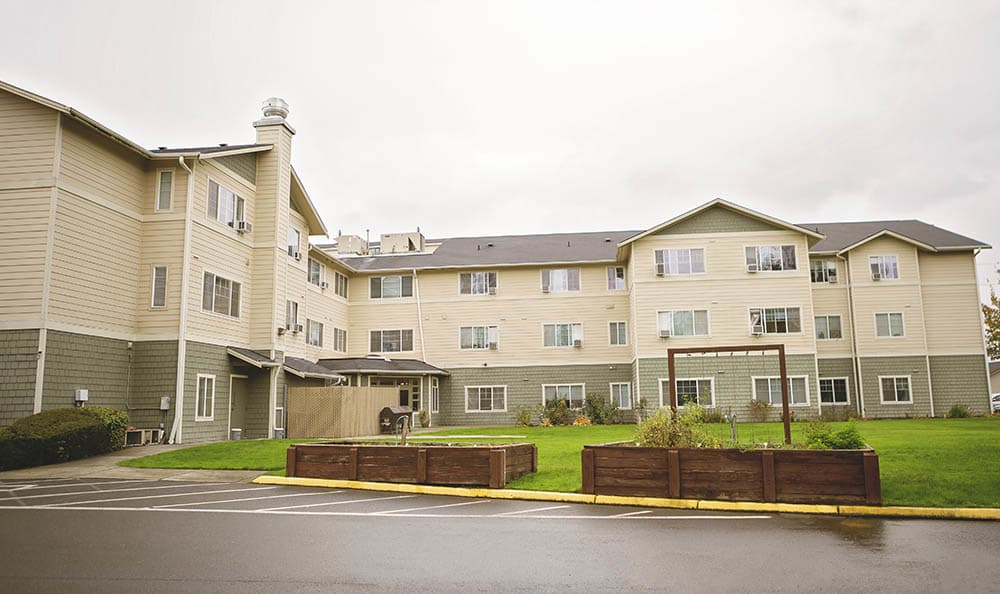 A residential apartment building with three stories, surrounded by a neatly manicured lawn under an overcast sky.