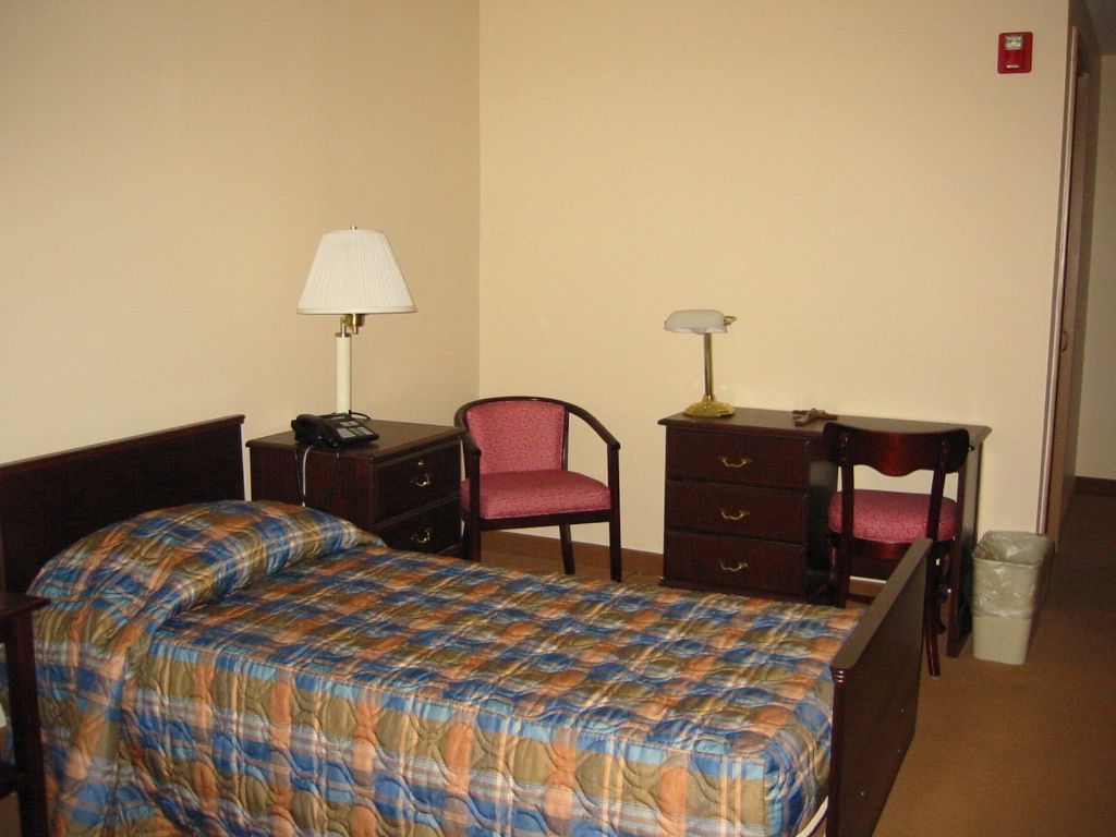 A simple bedroom with a bed, desk, chairs, and lamps.