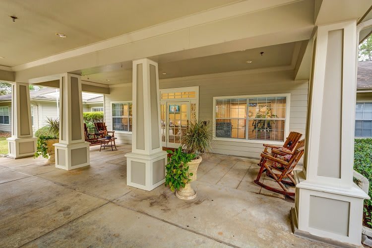 A covered patio area featuring rocking chairs, columns, and potted plants.