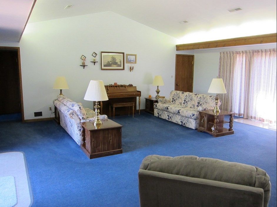 A vintage-style living room featuring blue carpeting, floral-patterned sofas, a piano, and multiple lamps.