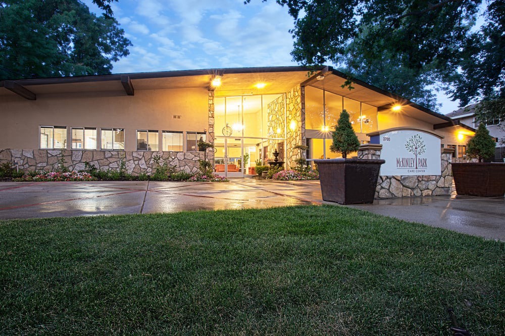 A modern building with illuminated windows during the evening, surrounded by well-maintained landscaping and trees.