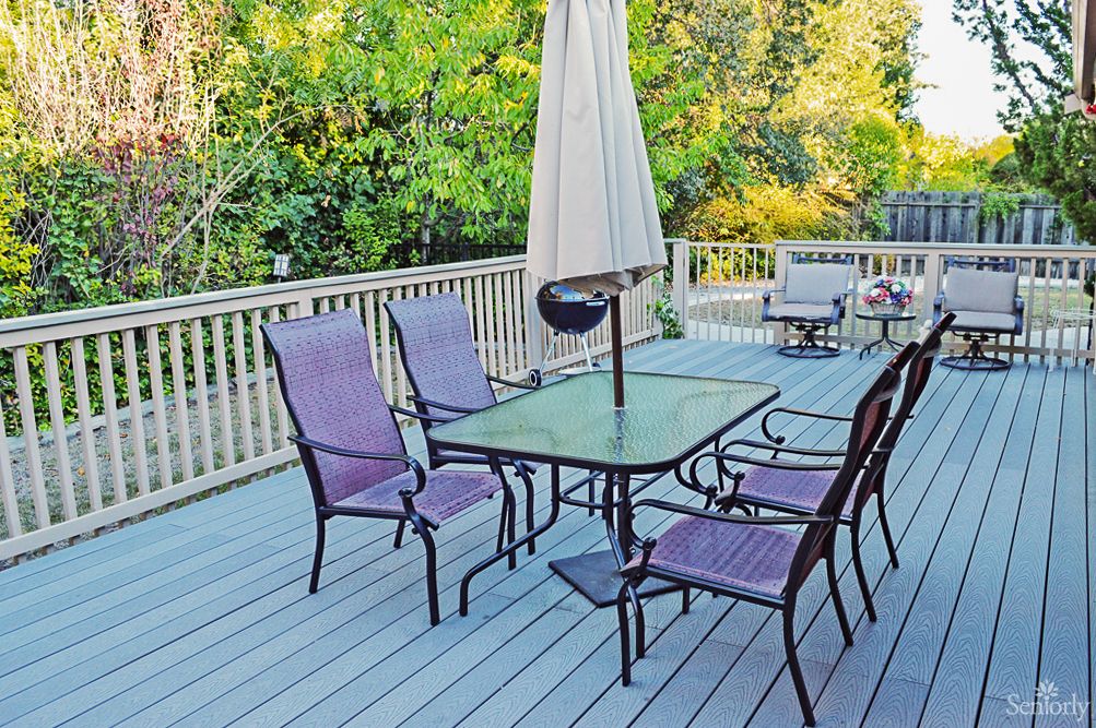 A cozy outdoor deck with patio furniture, including a glass table, chairs, and an umbrella surrounded by lush greenery.