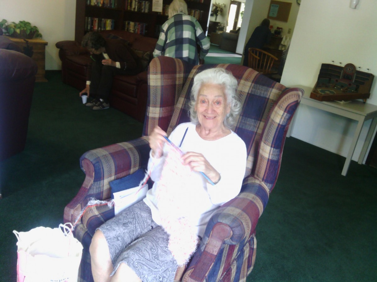 An elderly woman sits comfortably in an armchair while knitting in a cozy living room.