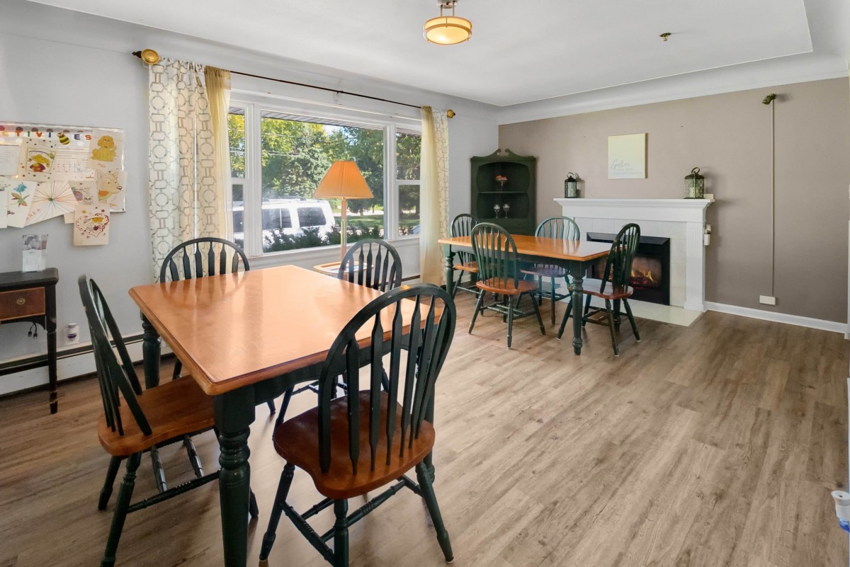 A cozy dining area with wooden tables and chairs, featuring a fireplace and large window with curtains.