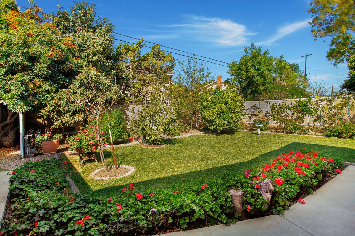 A beautifully landscaped backyard garden with green lawns, flowers, and trees under a clear blue sky.