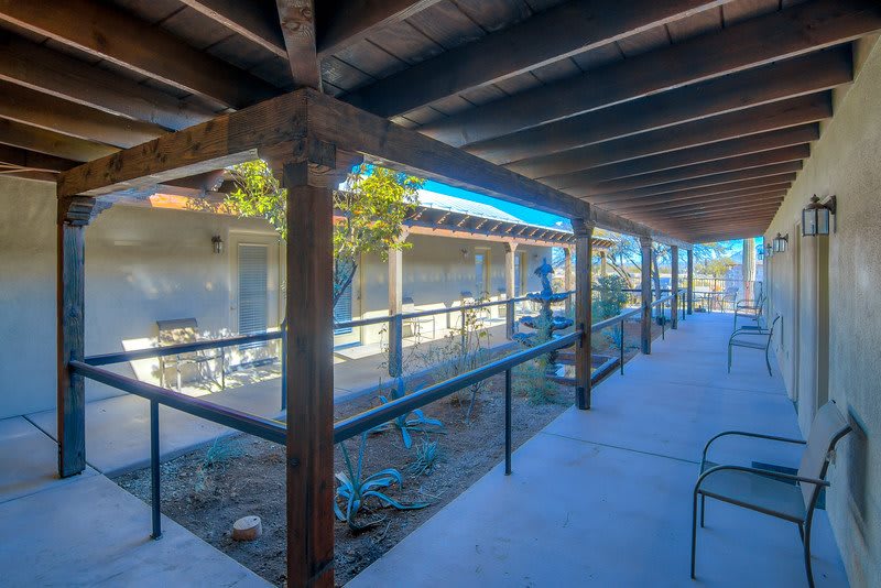 A sunny outdoor corridor with wooden beams and a ramp leading to an adjacent building.