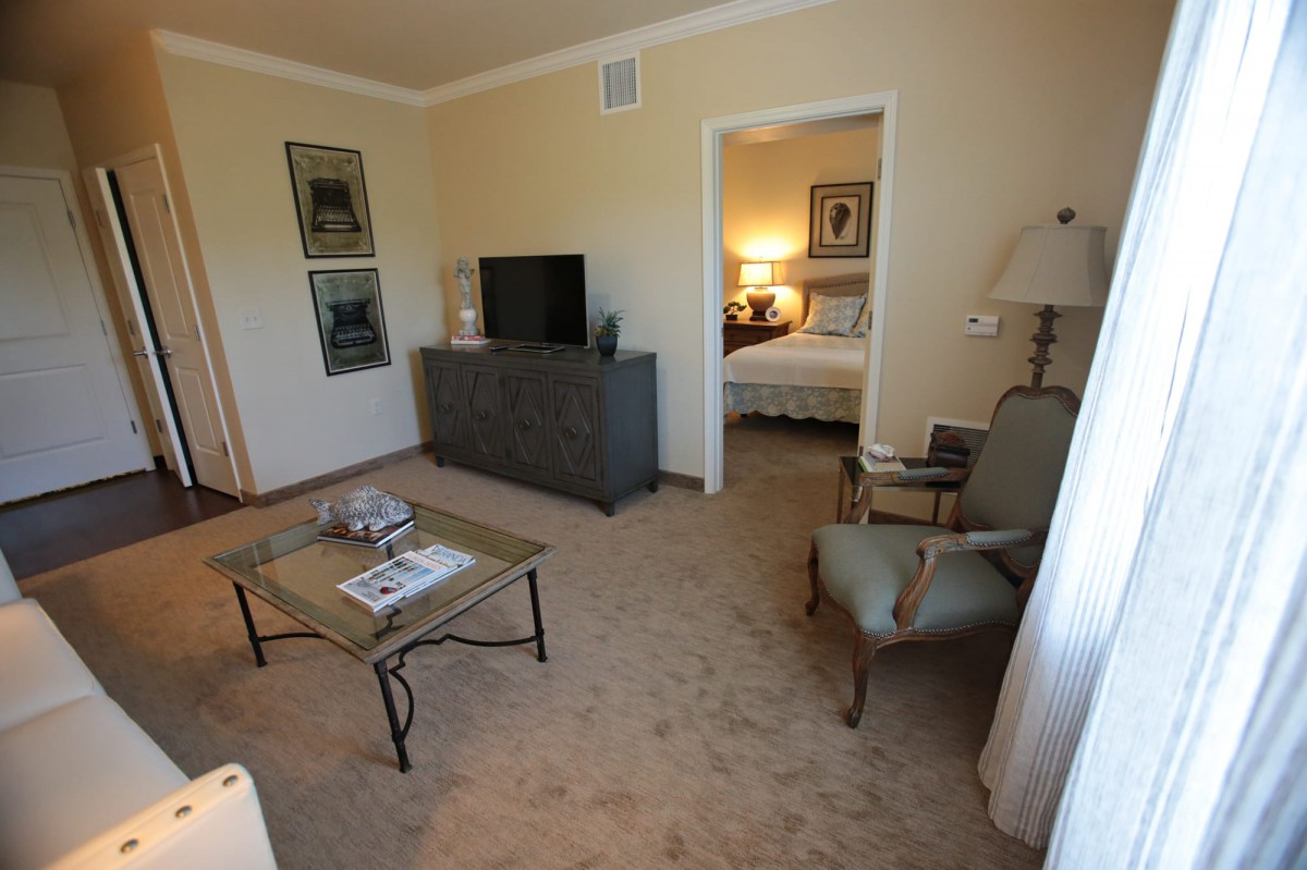 A cozy living room with a view into an adjoining bedroom, featuring a television, armchairs, and coffee table.