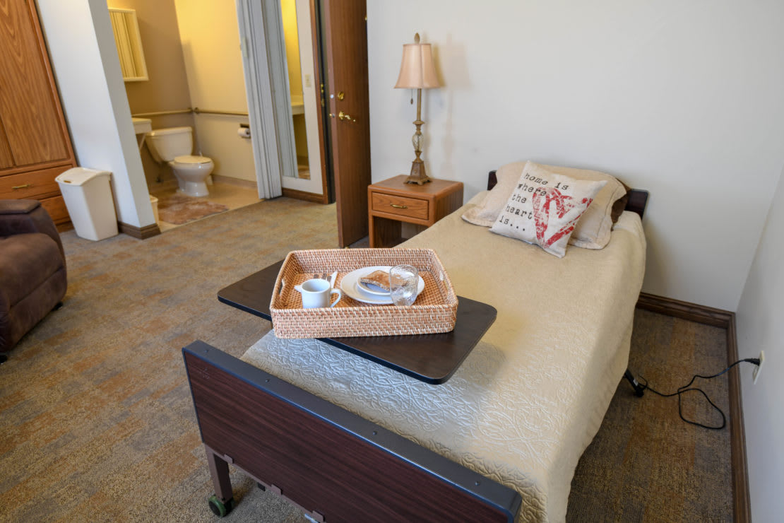 A cozy bedroom featuring a single hospital-style bed with a breakfast tray, a lamp on the nightstand, and an adjoining bathroom.
