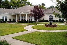 A charming house with a well-maintained lawn and a central pathway leading to the entrance, surrounded by lush greenery and a decorative fountain.