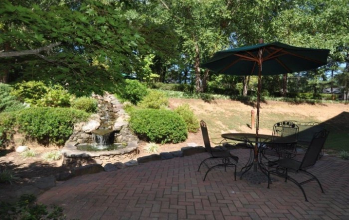 A serene garden patio with a fountain and outdoor seating under an umbrella.