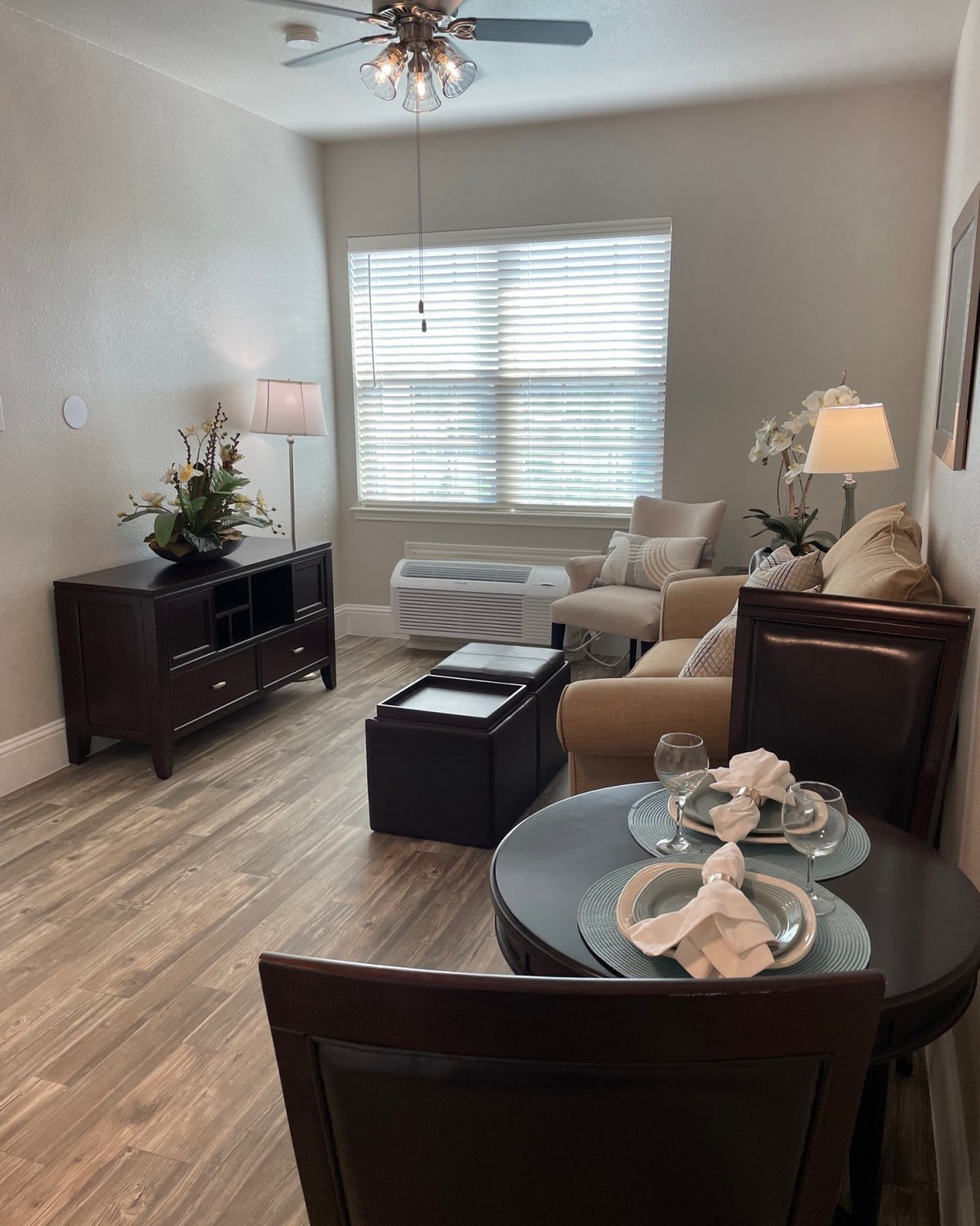 A cozy, modern living room with light wooden flooring, featuring a comfortable seating area and a small dining table near the window.