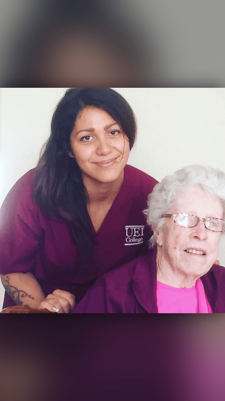 A caregiver in a nurse uniform poses with an elderly woman, both smiling.