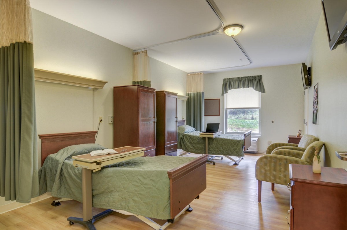 A hospital room featuring two beds, wardrobes, chairs, and a window with green curtains.
