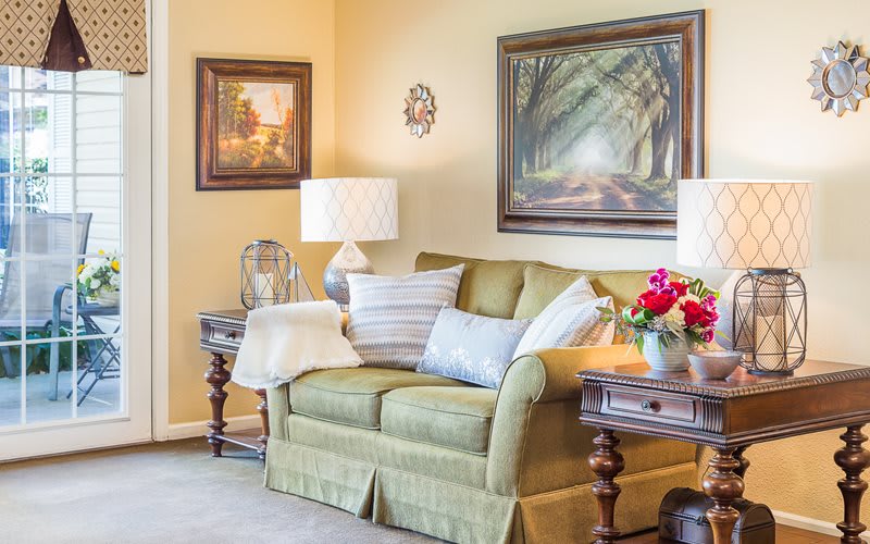 A cozy living room with a green sofa, adorned with cushions and flanked by wooden side tables with lamps. Paintings hang on the walls, adding warmth to the decor.