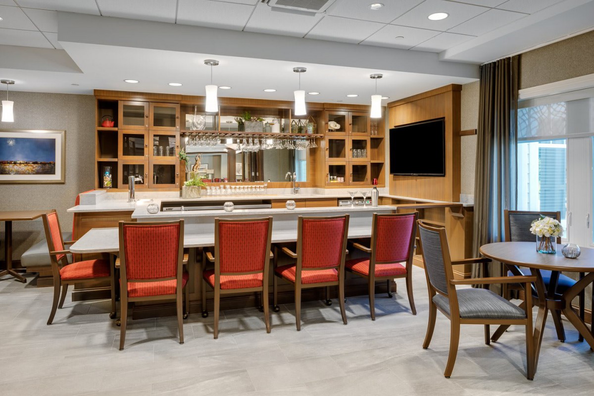 A modern bar area with red chairs and a well-organized counter, featuring elegant lighting and shelving.