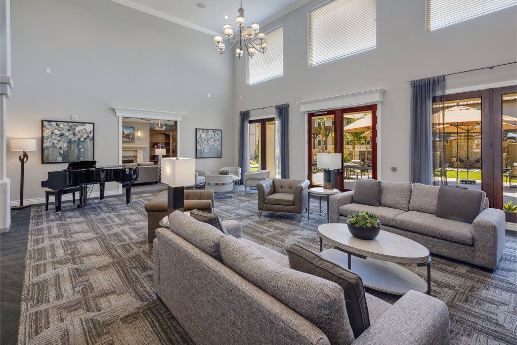 A spacious and elegantly designed living room featuring a modern seating area, grand piano, and chandelier under large windows.