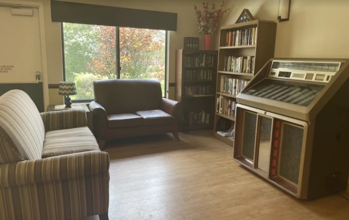 A cozy living room with comfortable seating, a jukebox, and bookshelves next to a large window.
