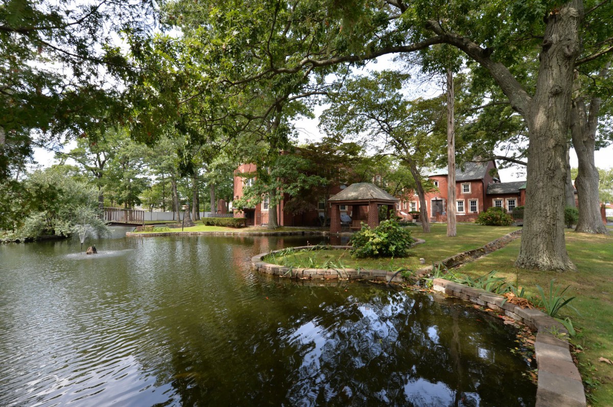 A tranquil scene of a brick building by a lakeside, surrounded by lush green trees and clear reflections in the water.