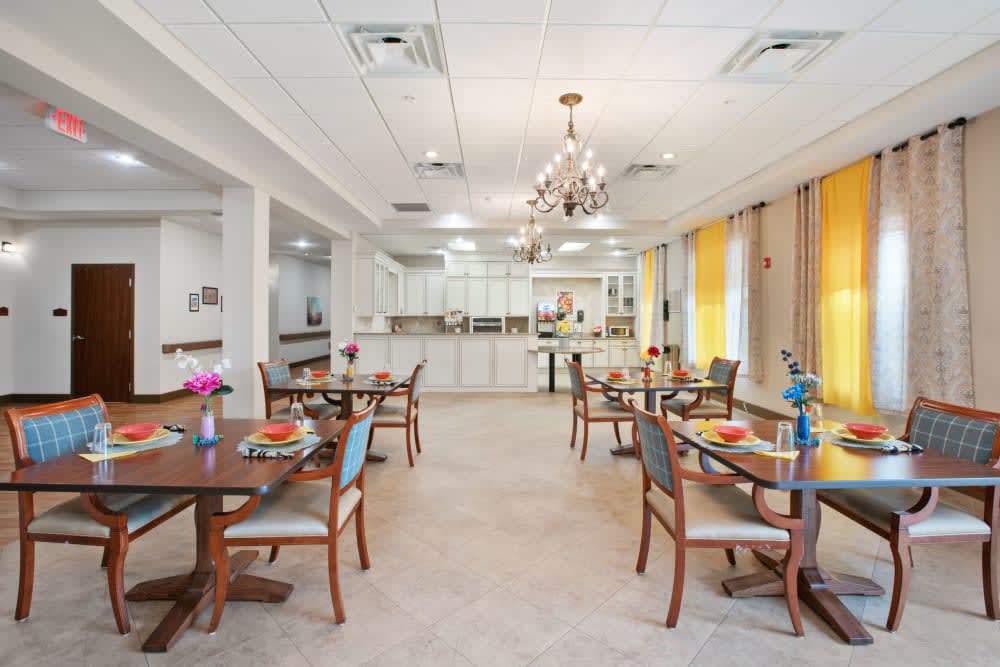 A well-lit dining room with neatly arranged tables and chairs, featuring a chandelier and adjacent kitchen area.