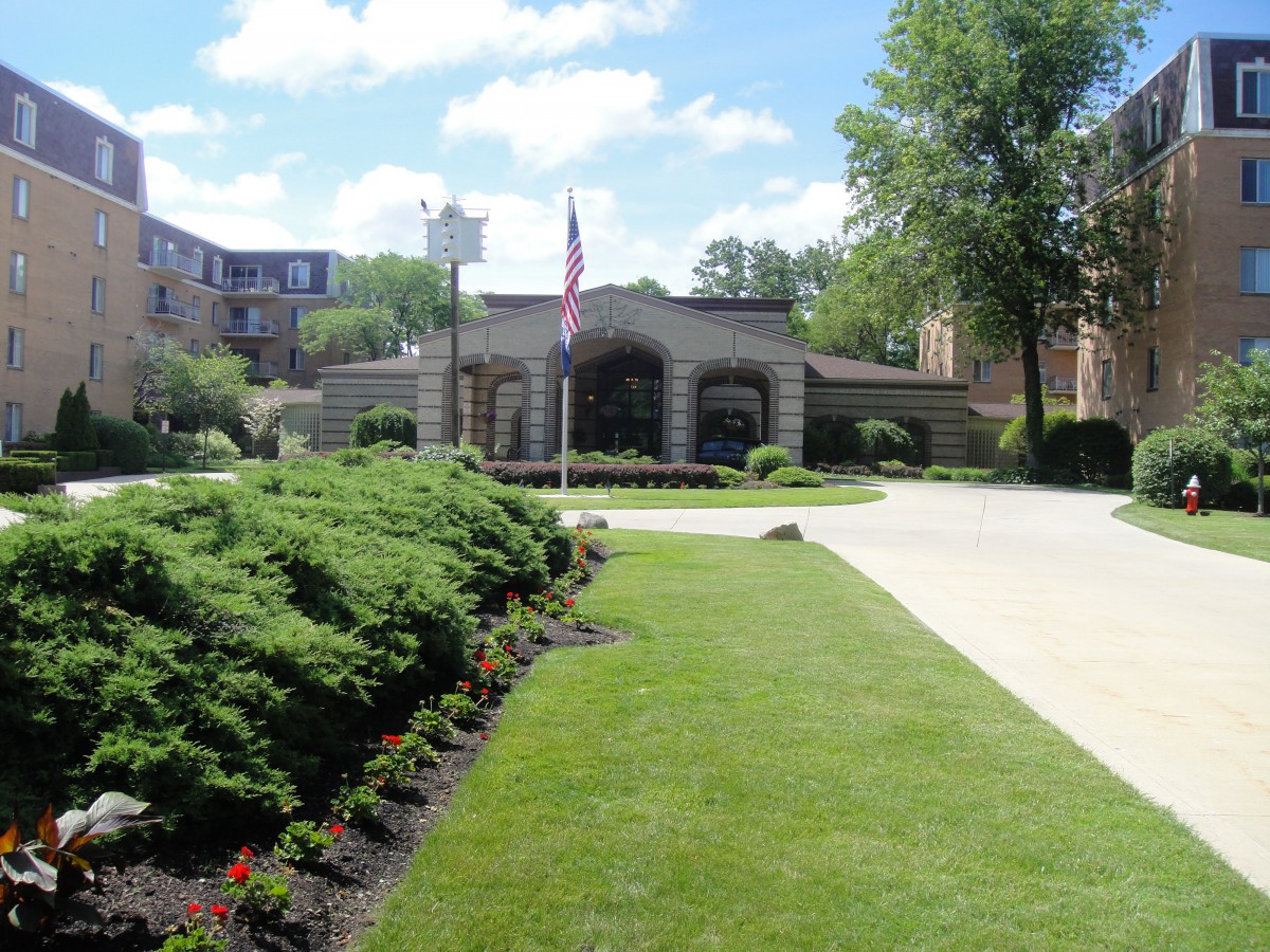 A landscaped entrance with a flag, surrounded by greenery and trees under a clear sky.