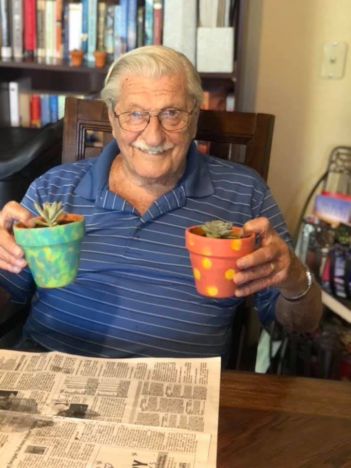 A smiling man holds two potted succulent plants indoors.