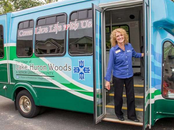 A smiling person stands at the open door of a bus, wearing a uniform.