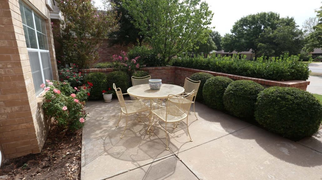 A cozy outdoor patio featuring a round table with four chairs, surrounded by lush greenery and neatly trimmed bushes.