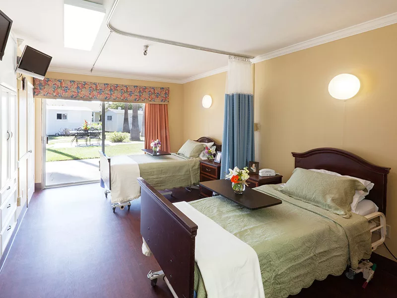 A well-lit hospital room with two patient beds and a view of an outdoor garden.