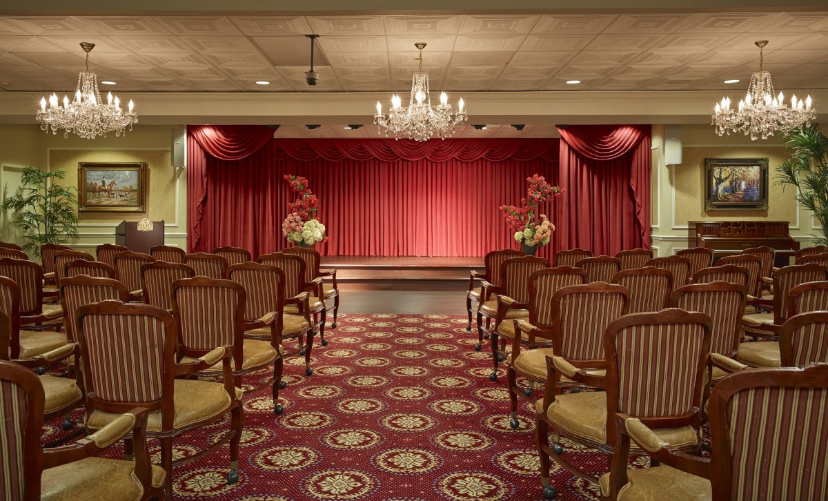 An elegant theater room with a stage set in front of red curtains, surrounded by rows of empty chairs and lit by a chandelier.