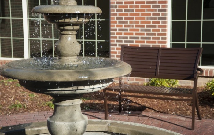 A stone fountain with flowing water next to a wooden bench, set against a brick wall outdoors.